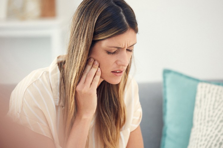 Woman in striped shirt on couch pressing hand to sore jaw