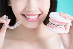 Nose down view of a woman smiling holding clear aligner in one hand and a model of teeth with braces in the other